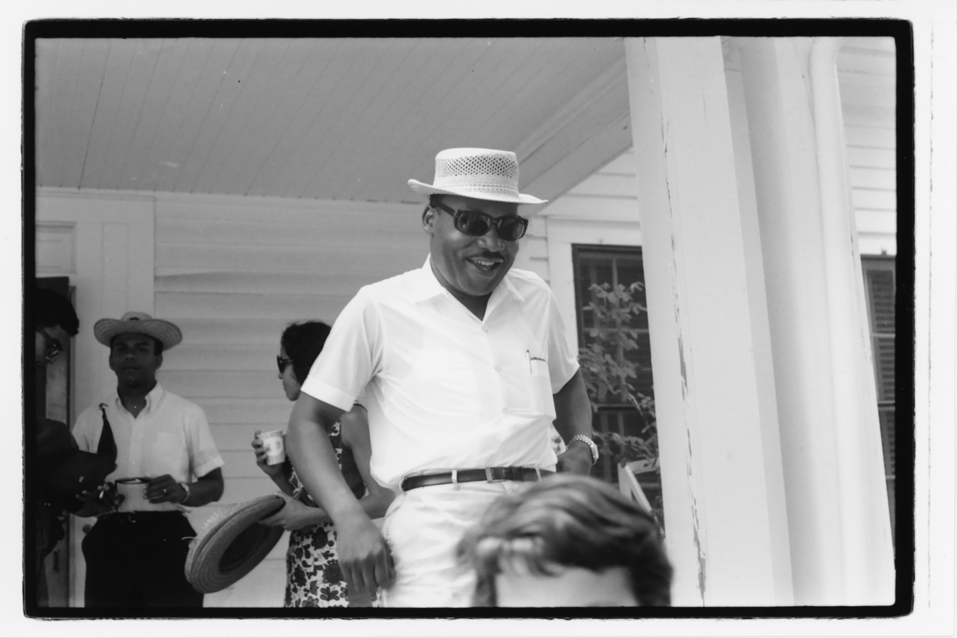Martin Luther King Junior, in a polo shirt, sunglasses, and boater hat, smiling from a porch at a summer gathering